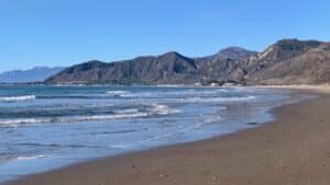This is a landscape scene of the beach, with ocean waves lapping at the shore, a broad expanse of sandy beach, and coastal mountains in the background. The sky is blue and clear. 